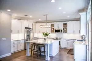 Modern kitchen with white cabinets, stainless steel appliances, a large island with two stools, a pendant light above, wood flooring, and a vase with green leaves on the island. Bright and spacious ambiance.