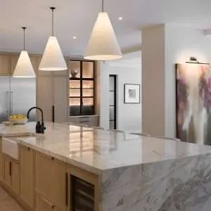 Modern kitchen with a large marble island, light wood cabinets, three cone-shaped pendant lights, and abstract art on the wall. A bowl of lemons sits on the countertop near a black faucet.