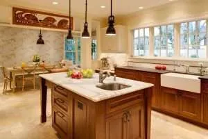 A spacious kitchen with wooden cabinets, a marble island countertop with a sink, pendant lights, and large windows showcases expert kitchen remodeling. A dining area with a floral wall and wooden table is visible in the background. Fruit bowls sit on the island.