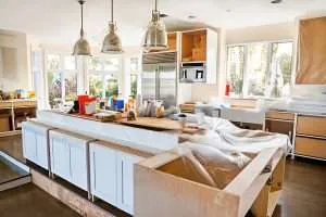 A kitchen under renovation with open cabinets, tools, supplies, and plastic coverings scattered across counters. Large windows let in natural light, and pendant lights hang above the unfinished island.