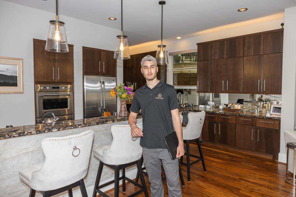 A man in a black shirt and cap stands in a modern kitchen with dark wood cabinets, marble countertops, and white barstools. He holds a tablet and faces the camera. Flowers and kitchen items are on the counters.