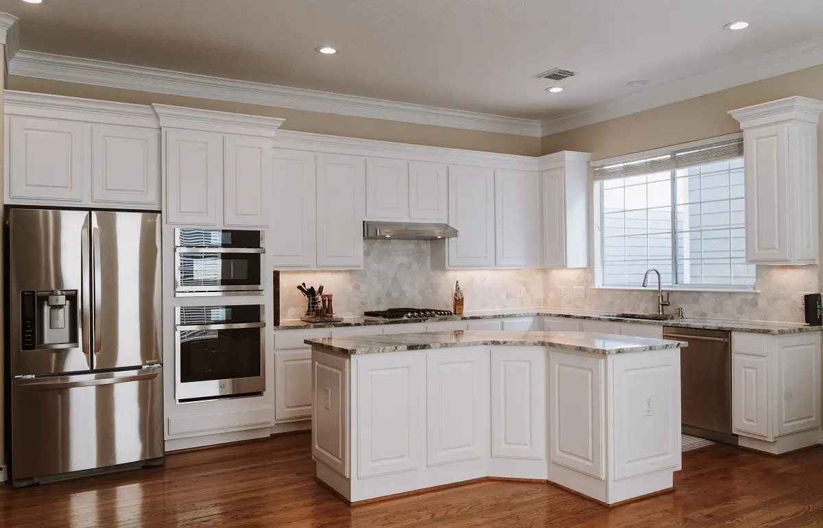 Modern kitchen with white cabinets, stainless steel appliances, marble countertops, double oven, refrigerator, dishwasher, and a stove under a range hood. Hardwood floors and a large window complete this stunning Kitchen Remodeling in Houston.