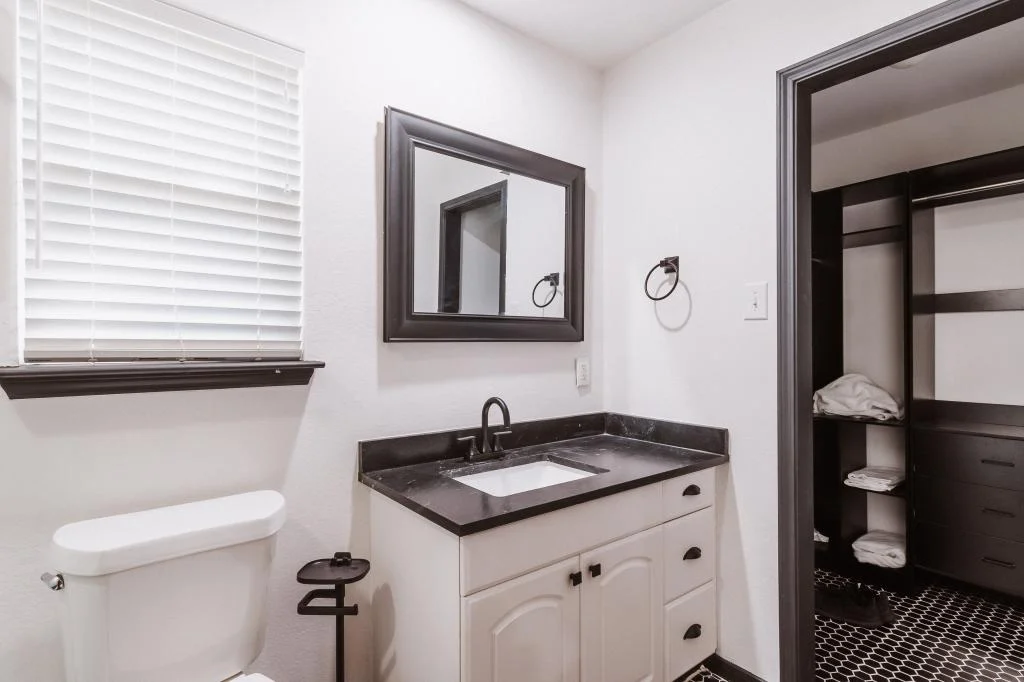 A modern bathroom remodeling Houston project featuring a white toilet, black countertop sink vanity, wall mirror, towel ring, and a walk-in closet with shelves and folded towels visible through an open doorway.