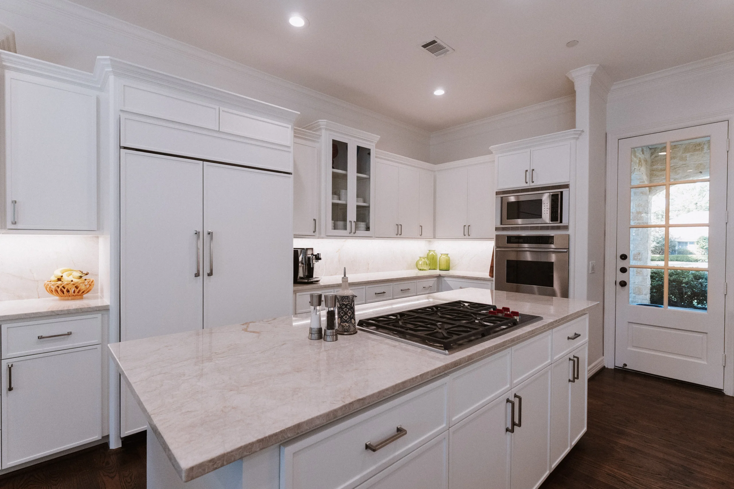 Modern kitchen with white cabinets, marble countertops, a large island with cooktop—perfect inspiration for kitchen remodeling Houston. Built-in appliances, under-cabinet lighting, and a glass door fill the space with natural light and style.