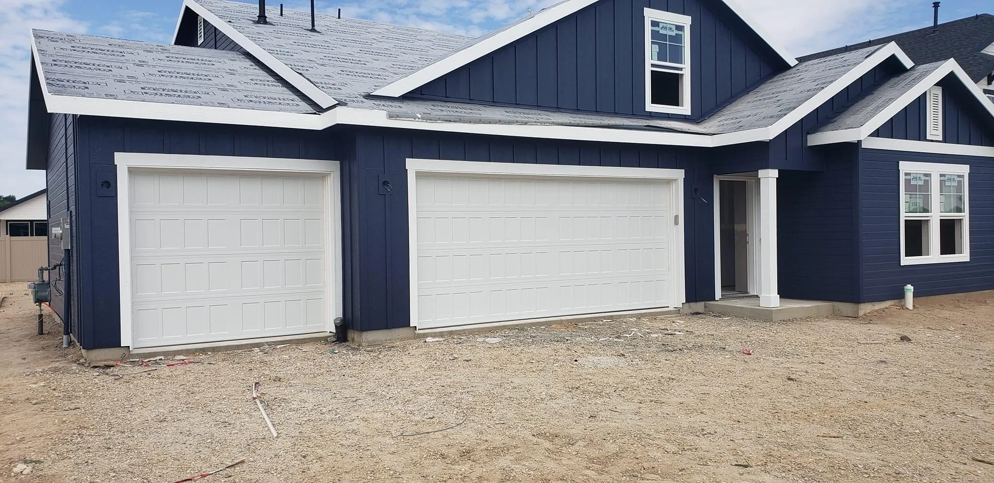 A newly built, dark blue house with white trim, three garage doors, and unfinished landscaping with bare dirt in front. The roof is partially covered with underlayment.
