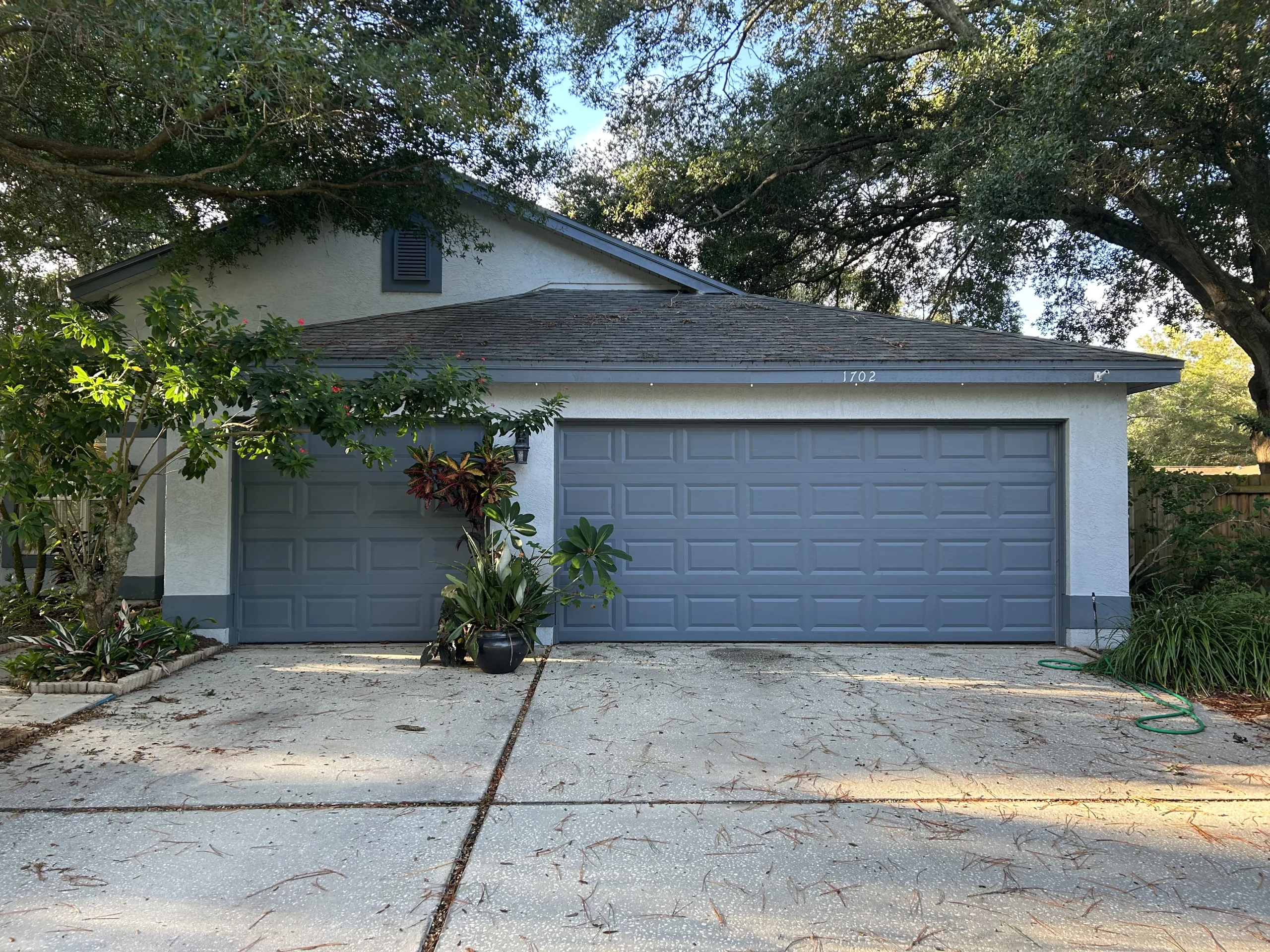 A house with two gray double garage doors, surrounded by trees and bushes. Potted plants are placed in front of the garages, and a green garden hose lies on the right side of the driveway.