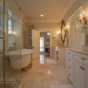 A spacious, elegant bathroom with a glass shower, freestanding bathtub, white cabinets, double sinks, large mirrors with gold frames, and soft lighting from wall sconces.