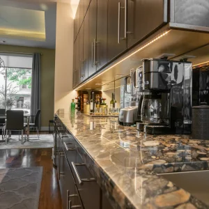 Modern kitchen with dark cabinets, a marble countertop, and various appliances including a coffee maker. Under-cabinet lighting highlights the workspace. Dining area with large windows is visible in the background.