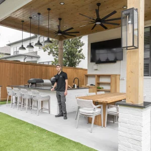 A man stands in a modern covered outdoor kitchen with a grill, dining area, bar seating, TV, ceiling fans, and wood accents, set on artificial grass in a fenced backyard.