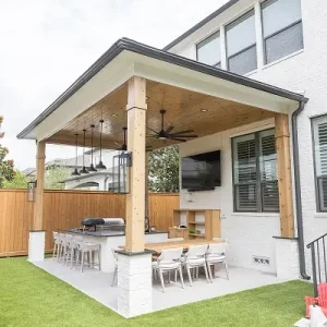 A modern backyard patio with a covered seating area featuring a long dining table, chairs, a TV, ceiling fans, pendant lights, a barbecue grill, and artificial grass lawn. The patio is attached to a white brick house.