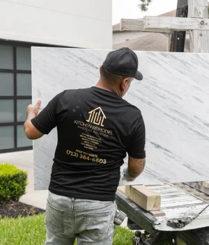 A worker in a black Kitchen Remodel shirt unloads a large marble slab from a truck outside a modern house, standing on grass beside a driveway.