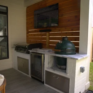 Outdoor kitchen setup featuring a grill, green ceramic smoker, mini fridge, and wall-mounted TV above a marble countertop—all designed by a top Kitchen Remodeling Company in Houston—set against a wooden privacy screen and fenced yard.