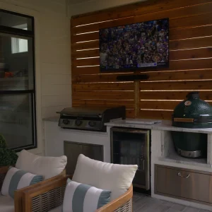 An outdoor kitchen features a grill, a Big Green Egg smoker, a mini fridge, and a wall-mounted TV displaying a sports game, with cushioned chairs in the foreground and a wooden privacy fence behind.