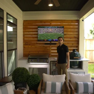 A man stands in an outdoor covered patio area with a grill, a TV displaying a football game, patio furniture, potted plants, and a green ceramic grill beside him. Sunlight comes in from the backyard.