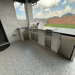 Outdoor kitchen with a stainless steel grill, side burner, sink, and mini fridge built into a white brick counter, set on a patterned tile patio with a view of a fenced backyard and blue sky with clouds.