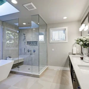 Modern bathroom with a glass-enclosed shower, freestanding bathtub, double-sink vanity with large mirror, white countertops, and a vase of flowers. Large windows and a skylight provide ample natural light.