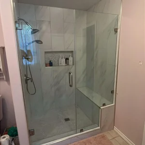Modern bathroom with a glass-enclosed shower featuring a built-in bench, wall-mounted showerhead, and shelves holding toiletries. A pink bath mat is on the tile floor outside the shower.