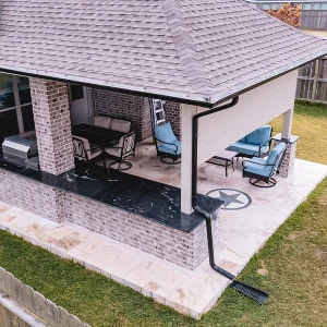 A covered backyard patio with outdoor furniture, a grill, and a black star decoration on the floor. The patio is attached to a brick house and surrounded by a fenced grassy yard.