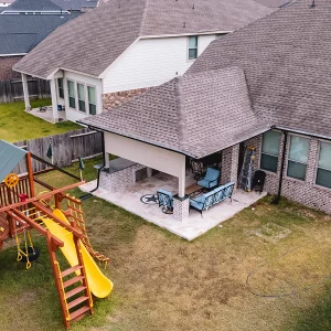 A fenced backyard with a wooden playset featuring slides and swings, next to a patio area with outdoor furniture behind a brick house with a covered porch.