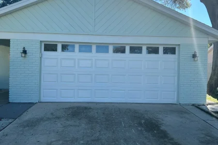 A white garage door with rectangular panels and small windows at the top is attached to a light-colored brick house—a great candidate for a Houston garage conversion or garage remodel. The concrete driveway leads past trees brightened by sunlight in the background.