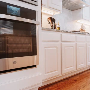 Modern kitchen with white cabinets, stainless steel built-in oven displaying the time, wooden floor, and various utensils and olive oil bottle on the countertop under bright lighting.