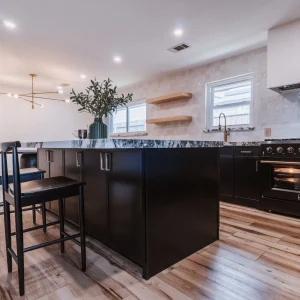 Modern kitchen with a black island, two black chairs, a marble countertop, oven, stovetop, open wooden shelves, hardwood floors, and decorative lighting. Large windows provide natural light.