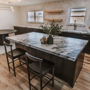 Modern kitchen with a large marble island, two black chairs, dark cabinets, wood flooring, and minimal decor. There are open shelves, two windows, and a vase with greenery on the island.