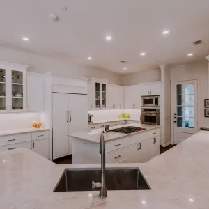 Spacious, modern kitchen with white cabinets, marble countertops, a central island with a sink, built-in stove, double oven, and a refrigerator. Recessed lighting brightens the room. A glass door leads outside.