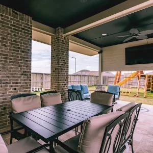 Covered patio with a black dining table and cushioned chairs, ceiling fan, mounted TV, and a view of a backyard with a wooden playset and blue outdoor seating. Brick walls and a fenced yard are visible.
