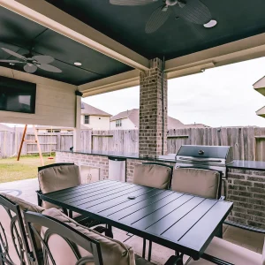 Covered outdoor patio with a black dining table, cushioned chairs, a TV mounted on the wall, ceiling fans, a barbecue grill, and a fenced backyard with a playset visible in the background.