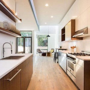 Modern kitchen with wooden cabinets, white countertops, stainless steel appliances, and open shelves with dishes. Large windows at the end let in natural light and reveal a sitting area and patio outside.