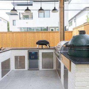Modern outdoor kitchen with white brick walls, black countertops, built-in grill, green ceramic smoker, mini fridge, sink, and wooden accents, set under a covered patio in a fenced backyard.