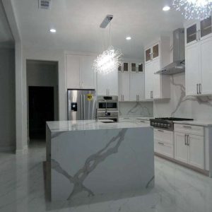 Modern kitchen with white cabinets, stainless steel appliances, marble-patterned island and countertops, built-in oven, and two contemporary pendant lights above the island.