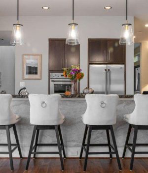 A modern kitchen with dark wood cabinets, stainless steel appliances, an island with a marble countertop, and four white upholstered barstools. Three pendant lights hang above the island.