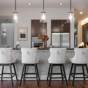 Modern kitchen with dark wood cabinets, stainless steel appliances, and a marble island. Four white upholstered barstools are lined up at the island, and pendant lights hang above it. A vase of flowers sits on the countertop.