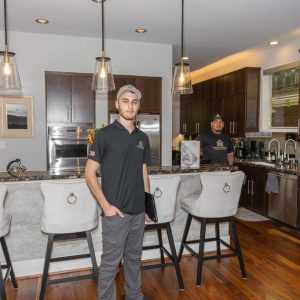 Two men stand in a modern kitchen with dark wood cabinets, marble countertops, pendant lights, and three upholstered bar stools at an island. One man stands in front, the other is near the sink in the background.