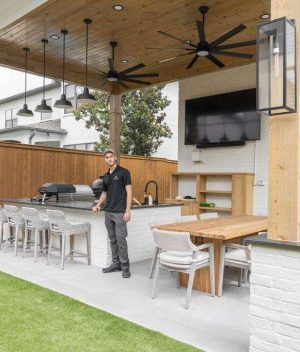 A man stands in a modern covered outdoor kitchen with bar seating, a dining table, a grill, a TV, ceiling fans, and wood accents, surrounded by a fenced yard with artificial grass.