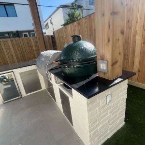 An outdoor kitchen with a built-in grill, a covered appliance, stainless steel cabinets, and a green ceramic kamado-style grill on a black countertop, set against a wooden fence.