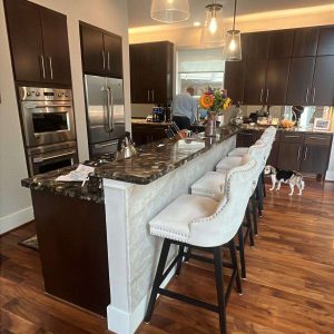 Modern kitchen remodel with dark cabinets, marble island countertop, three upholstered bar stools, pendant lights, stainless steel appliances, wooden floor, flowers on the island, and a dog in the background near the cabinets.