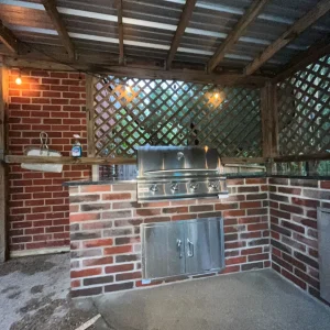 Outdoor kitchen area with a brick counter, built-in stainless steel grill, sink with soap dispenser, and lattice wood panels. Warm string lights hang above, creating a cozy atmosphere under a metal roof.