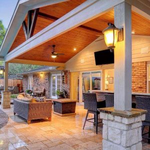 Covered outdoor patio with wooden ceiling, ceiling fans, and seating area next to a backyard pool. A bar with chairs lines the patio, and the space is warmly lit with lanterns. Lush greenery and a pergola are in the background.