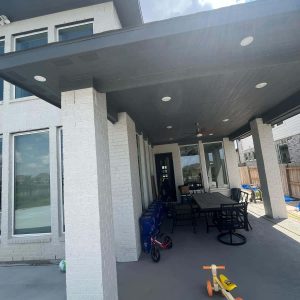 A covered patio attached to a modern white brick house features outdoor furniture and childrens toys, including a yellow tricycle, on the concrete floor. Recent countertop replacement in the home adds to its fresh, inviting feel under bright sunlight and blue sky.