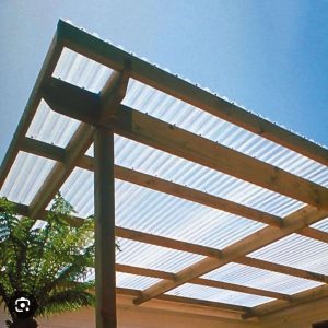 A clear corrugated plastic roof is supported by a wooden frame, providing shade while allowing sunlight through. A tree is partially visible beneath the structure, and the sky is clear and blue.