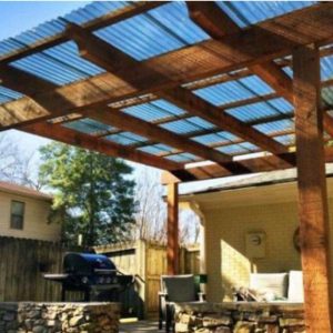 A backyard patio features a wooden pergola with a clear corrugated roof, outdoor seating, a stone wall, and a barbecue grill. The area is surrounded by a fence and neighboring houses.