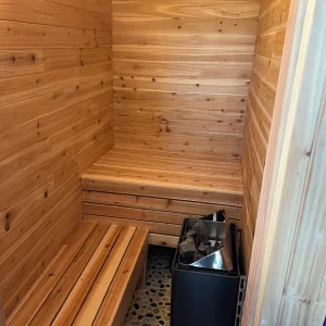 A small wooden sauna with two benches, a stone floor, and a metal stove with rocks in the corner. The walls and benches are made of light-colored wood.