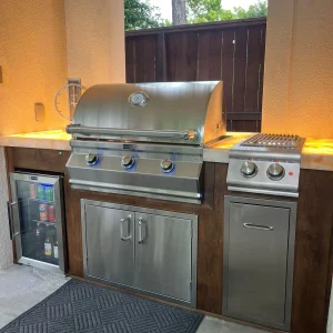 A built-in outdoor kitchen with a stainless steel gas grill, side burner, mini fridge, and storage cabinets, set against a stone patio and wooden fence with lights under the countertop.