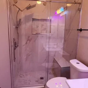 A modern bathroom with a glass-enclosed shower, a rainfall showerhead, white toilet, and a white vanity. The space features light wood-look flooring and marble-patterned tile walls, with colored light reflections visible.