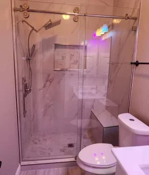 Modern bathroom with a glass-enclosed shower featuring a rainfall showerhead, built-in bench, and niche; a toilet and white vanity are also visible. Soft purple and yellow lights reflect on the surfaces.