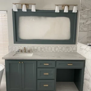 A modern bathroom vanity with dark green cabinets, gold hardware, a white countertop, a single sink, and a frosted rectangular mirror under two light fixtures. Walls have a marble and geometric tile design.