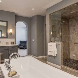 Modern bathroom with a large white bathtub, double sink vanity with mirrors, and a spacious glass-enclosed shower with beige tiles. Neutral gray walls and soft lighting create a calm, elegant atmosphere.